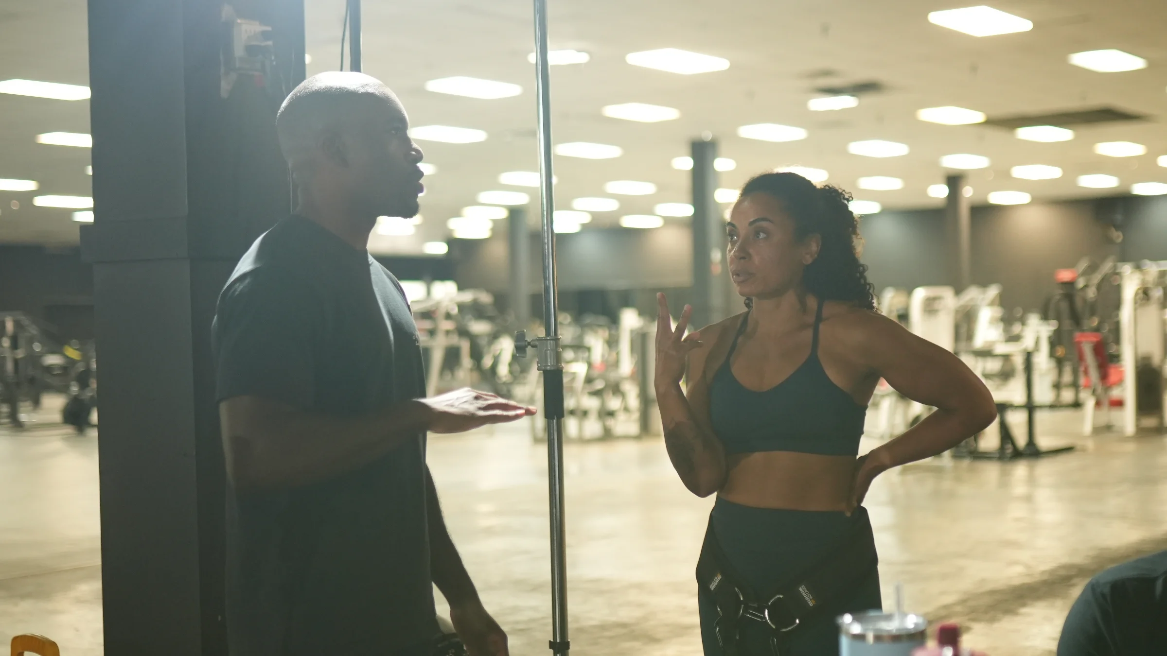 Tahirih speaks with her coach beside a squat rack in a gym, discussing her Streetlifting technique and competition preparation.